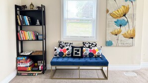 A room with a blue cushioned bench, a bookshelf with books and board games, a window with blinds, and a floral painting on the wall. - Ace Vacation Home at Niagara Falls - Canada (Niagara Falls)