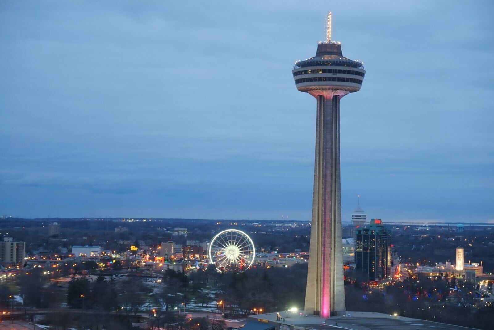 A tall tower with a circular observation deck, a Ferris wheel, and a cityscape with lights in the background.