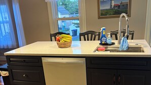 A kitchen with a central island, dark cabinetry, a dishwasher, and a sink with a faucet. There is a window with a view of the outside, a rug, and a painting on the wall. - Charming Beach Bungalow Near Everything. 3BR, 2 bath.  (Old Orchard Beach)