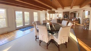 Un comedor espacioso con una gran mesa redonda y sillas blancas, vigas de madera en el techo y ventanas amplias con vistas al exterior.