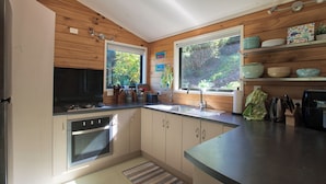 A modern kitchen with wooden walls, a stove, oven, sink, and a window overlooking greenery. - St Omer Hideaway: Love At First Sight (St Omer Bay)