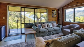 A cozy living room with a large window offering a scenic view, a grey sofa with patterned cushions, and a wooden ceiling. - St Omer Hideaway: Love At First Sight (St Omer Bay)