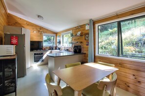 Een compacte keuken met een centraal eiland, een eethoek met tafel en stoelen, en grote ramen met uitzicht op de omliggende begroeiing.