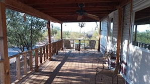 A wooden deck with a ceiling fan, a table with chairs, and a view of trees and cacti.