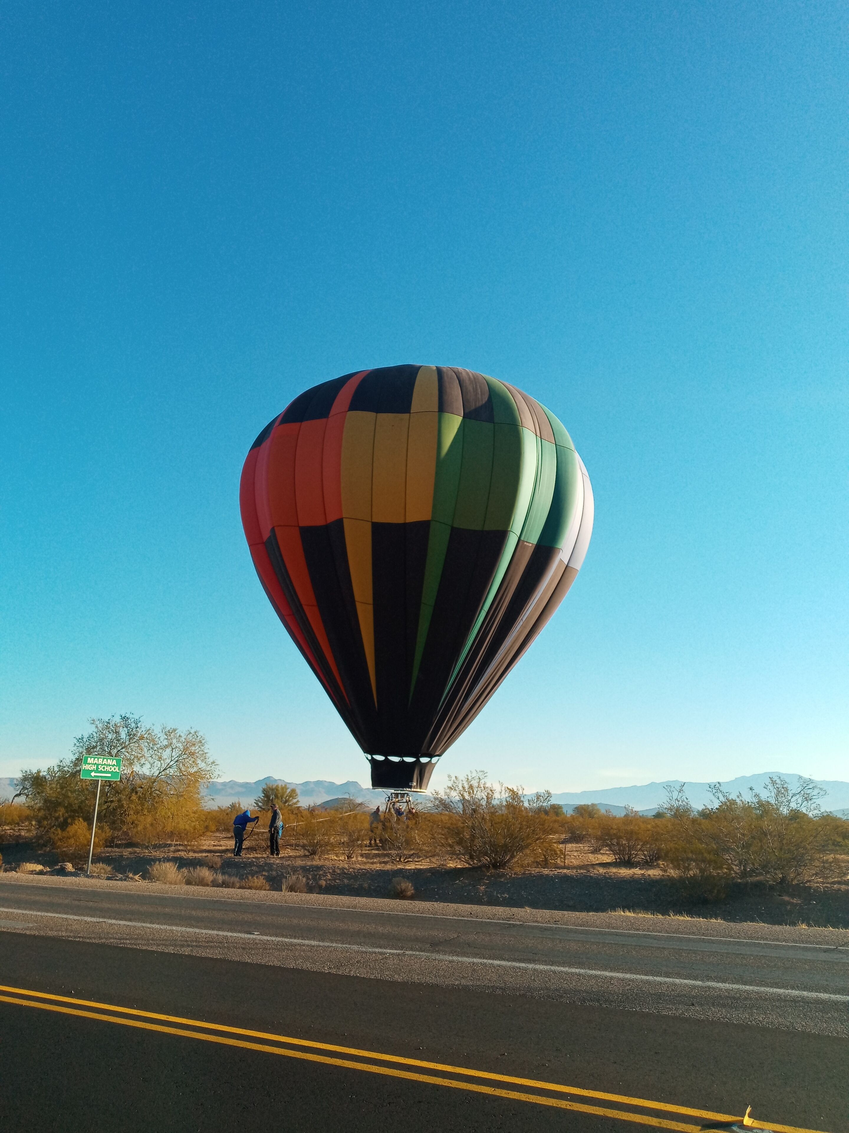 A hot air balloon with a multicolored design flying over a road.