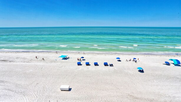 A beach with white sand and clear turquoise water. There are several blue umbrellas and lounge chairs.