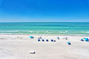 A beach with white sand and clear turquoise water. There are several blue umbrellas and lounge chairs.