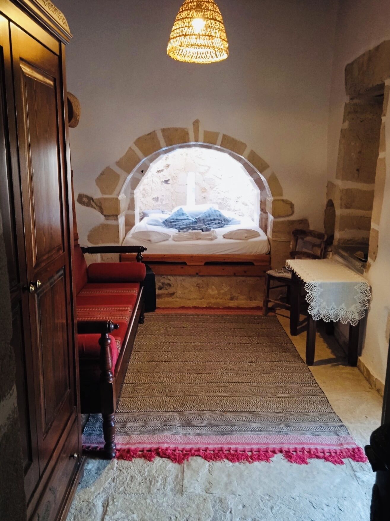 A cozy room with a stone fireplace, a wooden wardrobe, a bed with a red headboard, and a hanging light fixture.