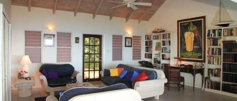 A living room with a high ceiling, a ceiling fan, a sofa set, a coffee table, a bookshelf, and a door with shutters.