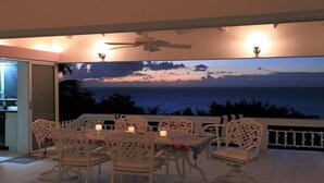 Un comedor con una mesa dispuesta para seis personas, rodeada de sillas blancas con diseños elaborados en el respaldo. Se observa un ventilador de techo y la habitación cuenta con un amplio ventanal con vista al océano al atardecer.