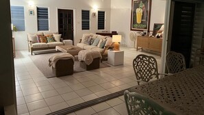 Una sala de estar con un sofá, dos otomanas, una mesa de centro y una mesa de comedor con sillas. Hay un ventilador y un cartel en la pared.