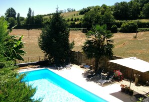 Une piscine entourée d’une clôture et de verdure, avec des chaises longues et une table à proximité.
