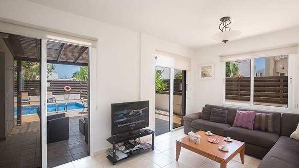 Una sala de estar con televisor, sofá, mesa de centro y una piscina visible a través de puertas de cristal.