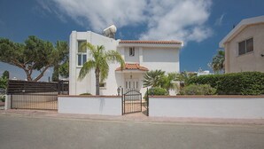 Una casa moderna de dos pisos con fachada blanca y techo de tejas rojas, acceso por puerta enrejada y un jardín cuidado con palmeras.