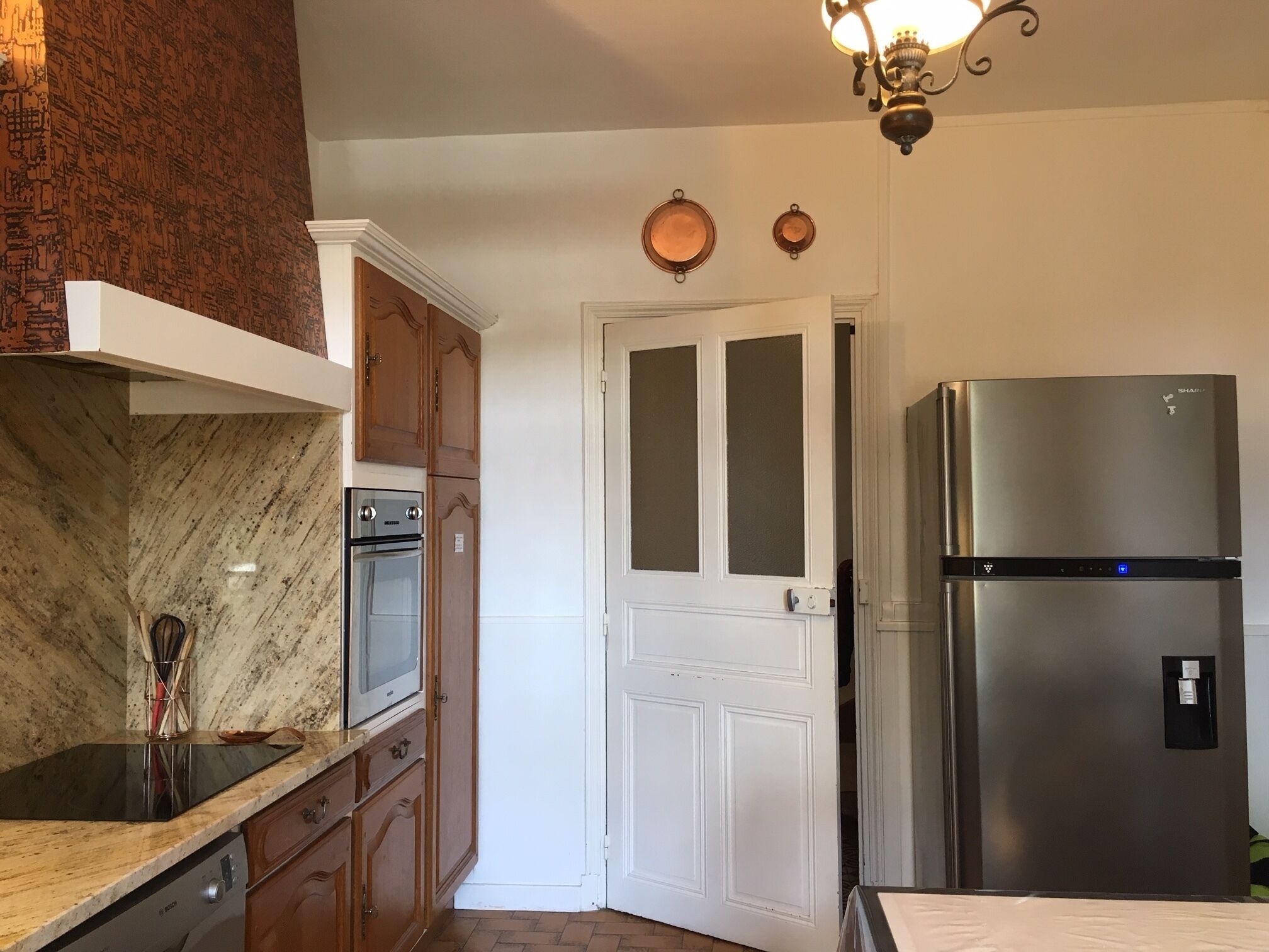 A kitchen with a stainless steel refrigerator, a built-in oven, wooden cabinets, a marble countertop, and a white door with frosted glass panels.