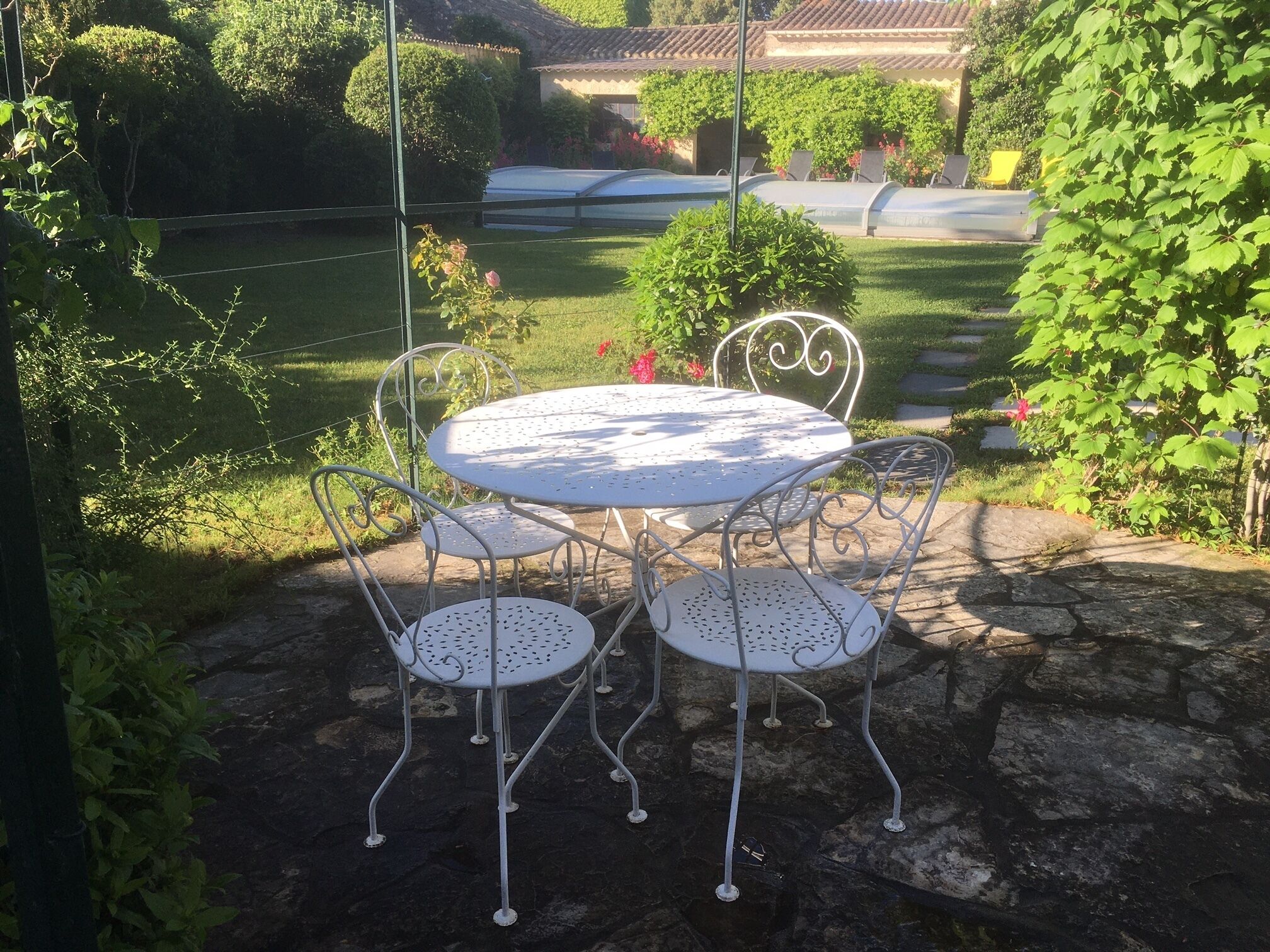 Um pátio com uma mesa redonda e quatro cadeiras, cercado por pavimento de pedra e vegetação.