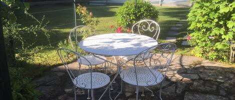 Um pátio com uma mesa redonda e quatro cadeiras, cercado por pavimento de pedra e vegetação.