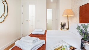 Une chambre d’hôtel avec un lit, une table de chevet avec une lampe, un magazine sur le lit et une porte donnant sur une autre pièce.