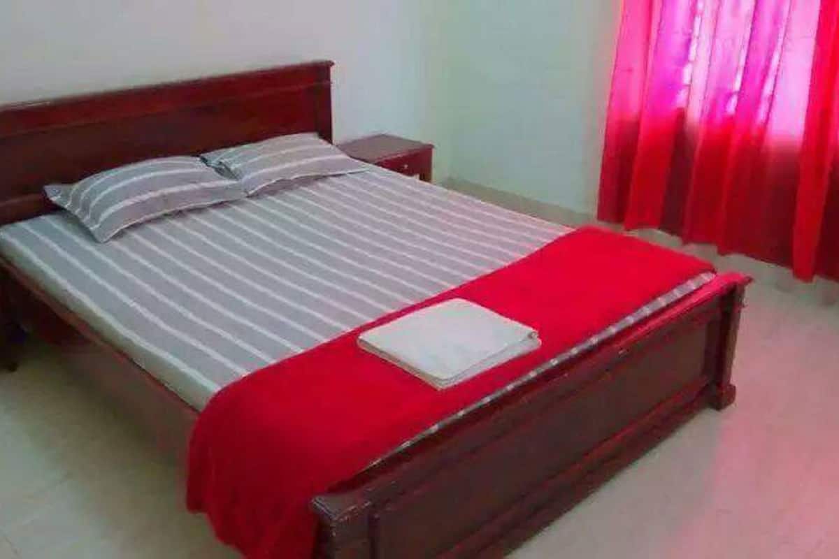 A single bed with a wooden headboard, a red bedspread, and a white pillow. There is a nightstand with a lamp and a book on it.