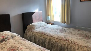 Un dormitorio con dos camas, una mesita de noche con lámpara, una ventana con cortinas y un cuadro enmarcado en la pared.