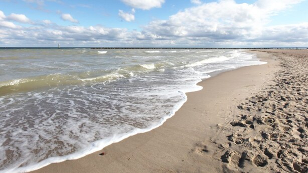 Una spiaggia con onde che lambiscono dolcemente la riva, un cielo sereno e un molo in lontananza.