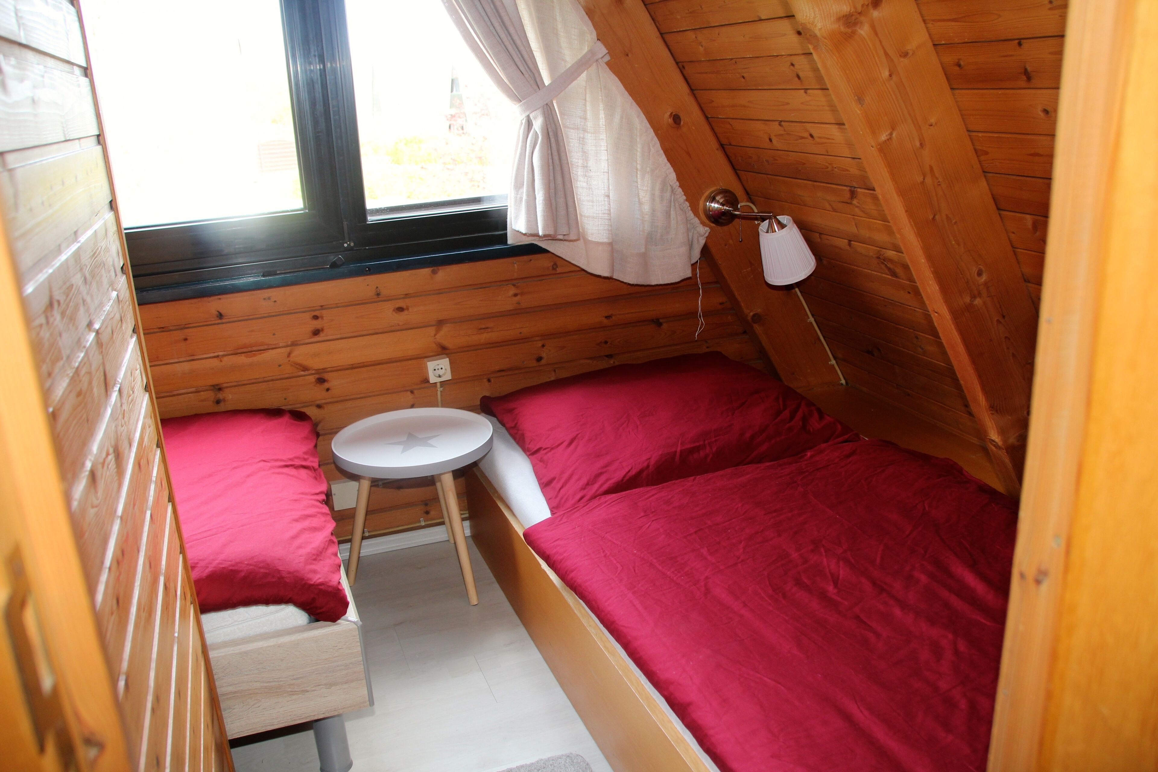 A small, cozy room with a wooden sloped ceiling, a single bed with red bedding, a small round table, and a window with curtains.