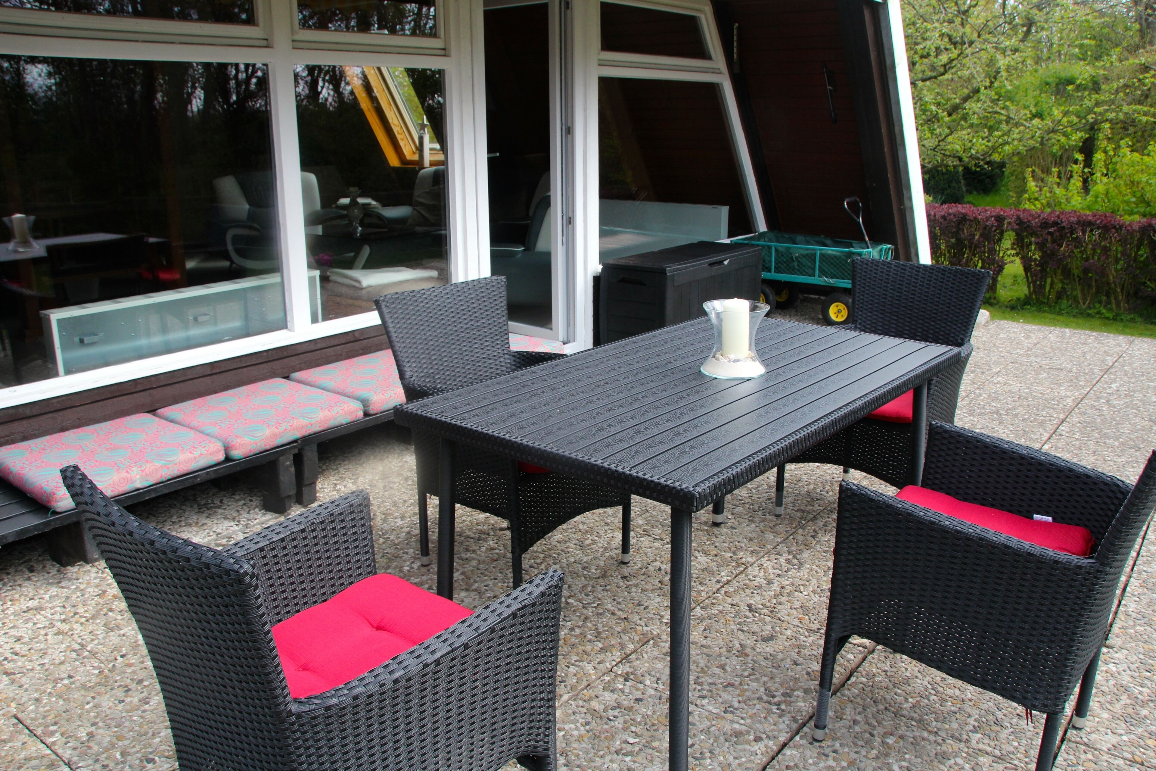 Une terrasse extérieure avec une table et quatre chaises, un banc avec des coussins rouges et un chariot vert.
