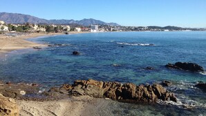 A coastal beach with clear blue water, rocky shoreline, and a cityscape in the background. - Apartment Ocean Front - Rocas del Mar - Riviera del Sol. (Mijas Costa)