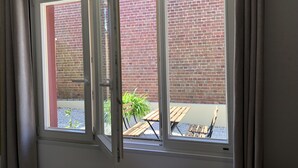 Una habitación con ventana que da a un muro de ladrillo y un patio con mobiliario de madera.