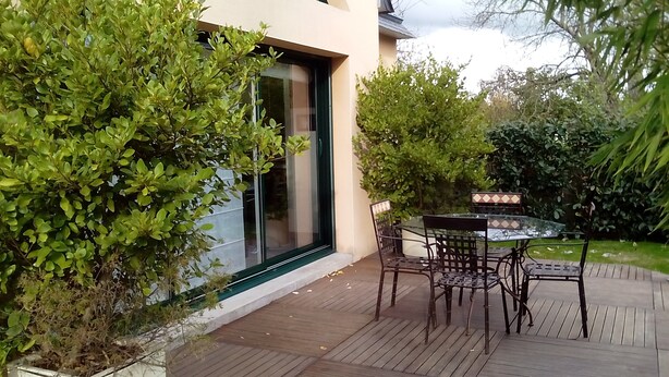 A patio area with a glass table and chairs, surrounded by greenery and a modern building facade. - HOUSE QUIET, COMFORTABLE, LUMINOUS (Cesson-Sévigné)