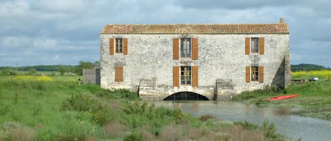 Edificio de piedra con tejado de tejas y contraventanas de madera, ubicado junto a un río tranquilo y rodeado de vegetación.