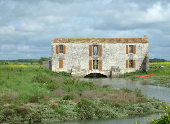 A stone building with a tiled roof and wooden shutters, situated by a calm river with surrounding greenery.