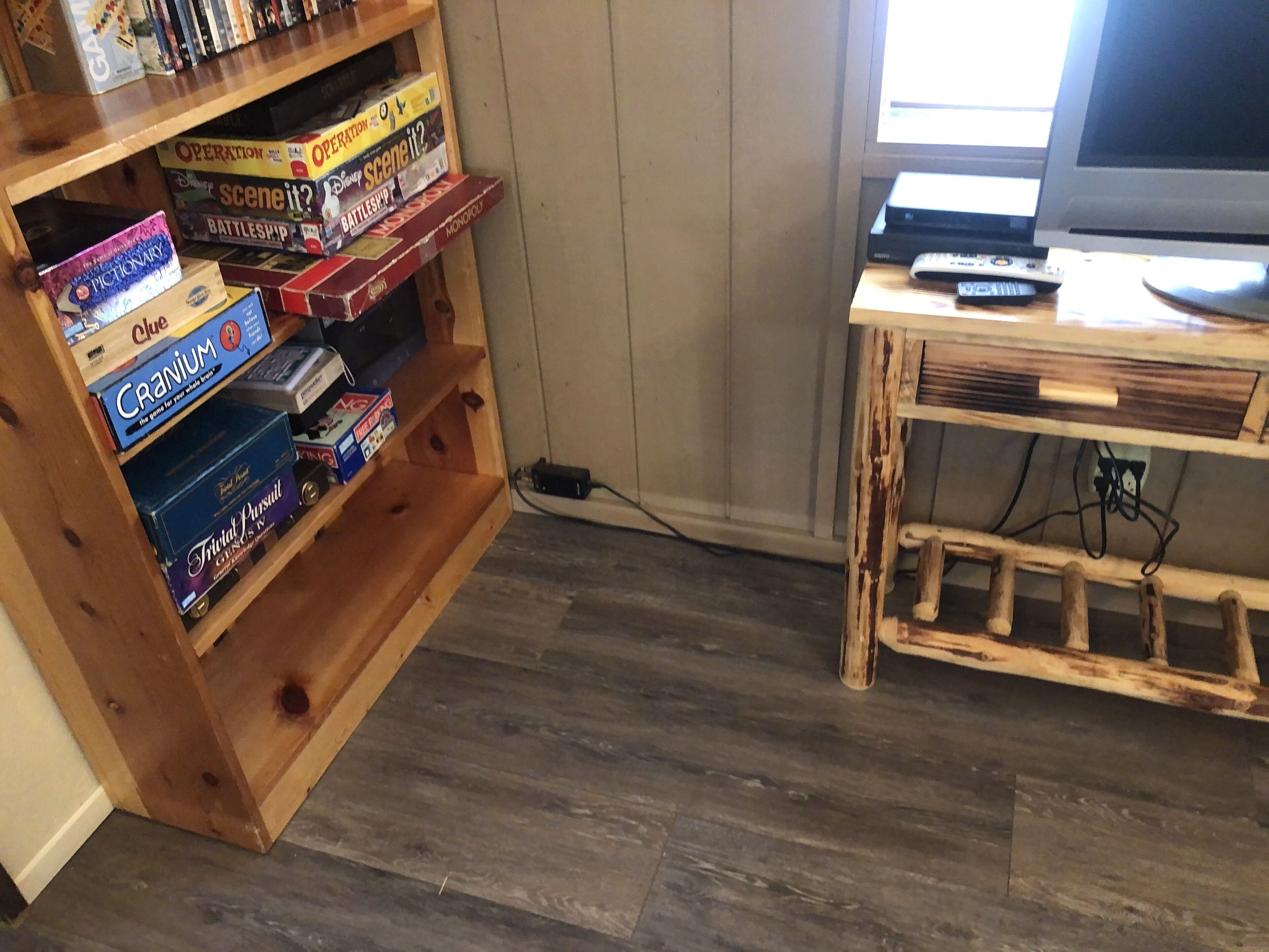 A room with a wooden bookshelf filled with board games, a wooden TV stand with a remote, and a wooden floor.