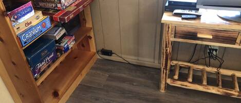A room with a wooden bookshelf filled with board games, a wooden TV stand with a remote, and a wooden floor.