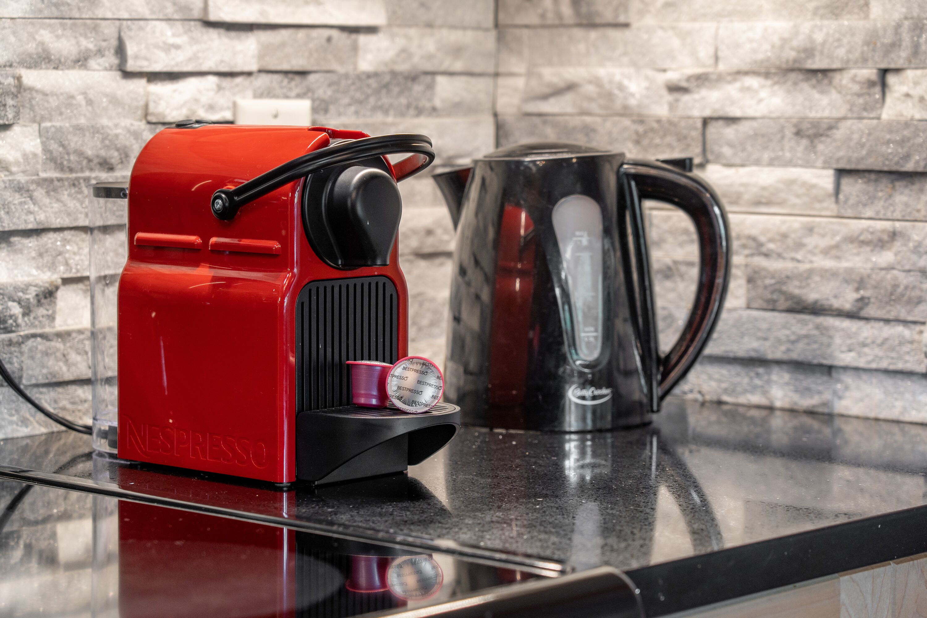 A red Nespresso machine and a black coffee pot on a countertop with a stone wall background.