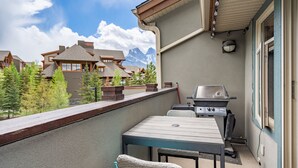 A balcony with a grill, a table, and chairs, overlooking a mountainous landscape. - Canmore Penthouse Loft / Round  Heated Outdoor Pool (Canmore)