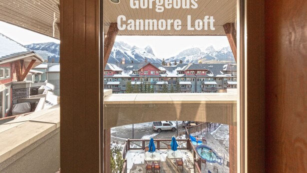 A view from a hotel window labeled "Gorgeous Canmore Loft" with a mountain backdrop and a pool area visible below. - Canmore Penthouse Loft / Round  Heated Outdoor Pool (Canmore)
