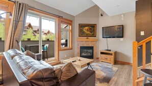 A modern living room with a leather sofa, a fireplace, a flat-screen TV, and a large window with curtains. - Canmore Penthouse Loft / Round  Heated Outdoor Pool (Canmore)