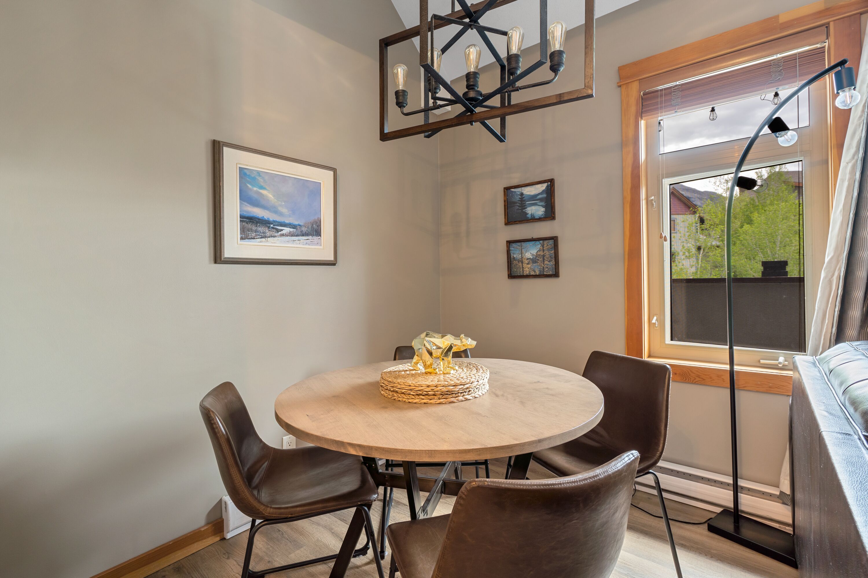 A dining area with a round table, four chairs, and a chandelier. There are framed pictures on the wall and a window with a view of greenery.