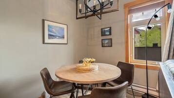 A dining area with a round table, four chairs, and a chandelier. There are framed pictures on the wall and a window with a view of greenery.