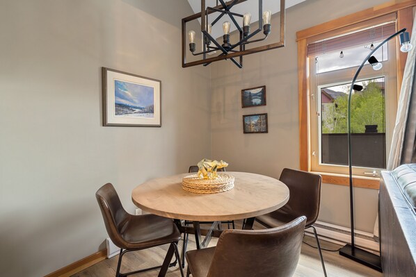 Un coin repas avec une table ronde, quatre chaises et un lustre. Les murs sont ornés de tableaux encadrés, et une fenêtre offre une vue sur la verdure.