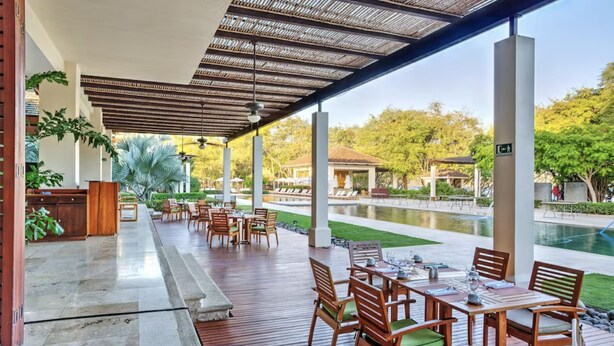 Un espace extérieur abrité, aménagé pour les repas, avec des tables et des chaises en bois, une piscine en arrière-plan et une vue sur la verdure environnante.