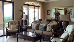 Un salon spacieux comprenant un grand canapé, deux fauteuils et une table basse. Une grande fenêtre offre une vue sur la verdure environnante.