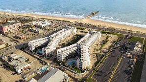 Vue aérienne d’un complexe hôtelier situé sur la côte, composé de plusieurs bâtiments, d’un parking et d’une plage en arrière-plan.