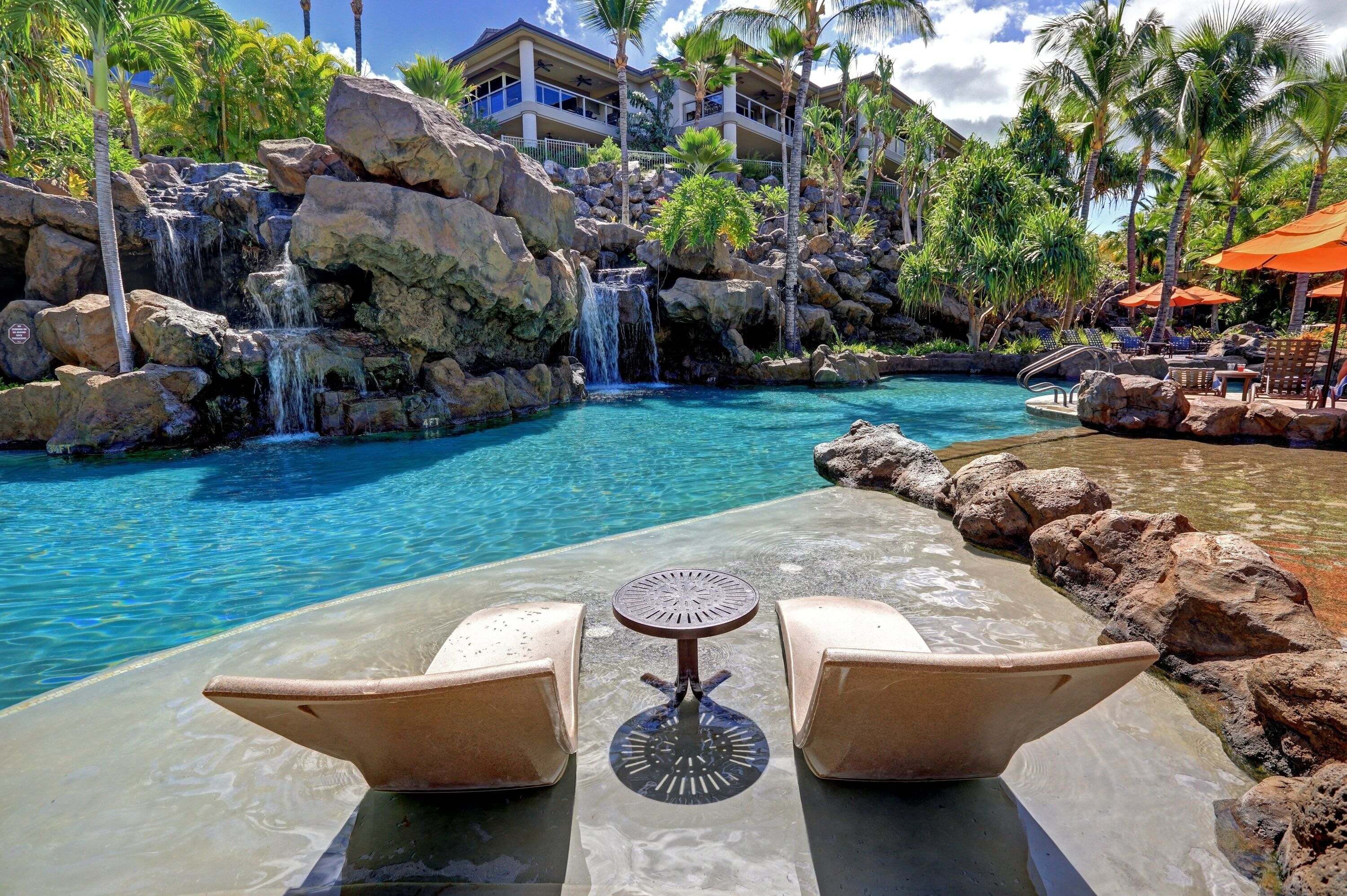 A poolside area with a waterfall, lounge chairs, and a view of a resort building.