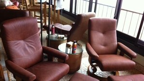 Een kamer met twee bruine leren fauteuils en een bijpassend voetenbankje, een klein rond tafeltje met een glazen blad, en uitzicht buiten door een raam.