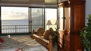 Een slaapkamer met een bed, stoelen, een houten kast en uitzicht op zee.