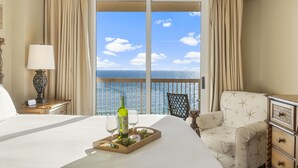 A hotel room with a bed, a nightstand with a lamp, a chair, and a view of the sea through a large window.