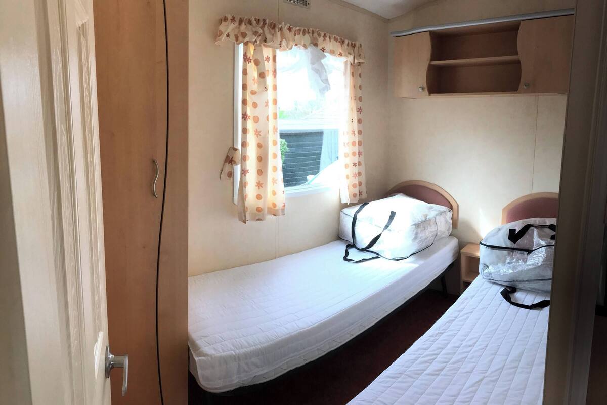 A small, single-bed room with a window, a curtain, a bedside table, and a shelf above the bed.