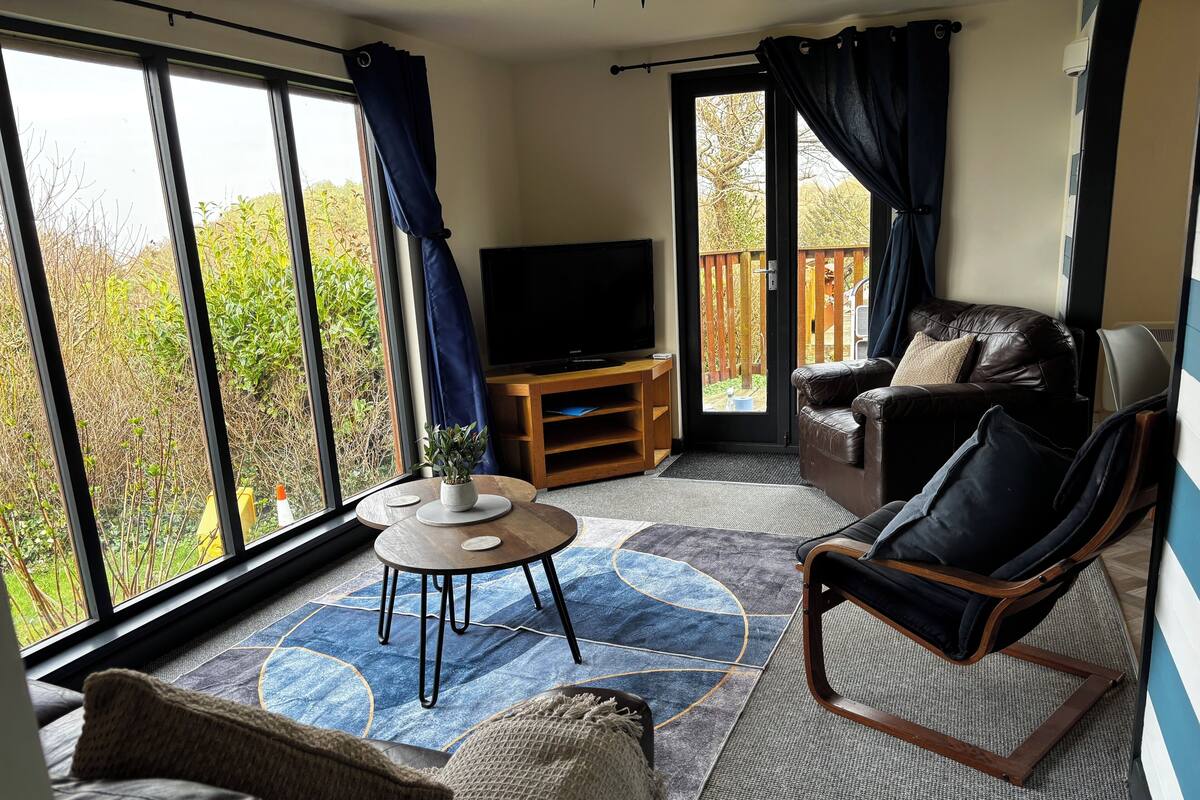 A spacious living area with a large window, a flat-screen TV, a wooden entertainment unit, a leather sofa, and a wicker armchair.